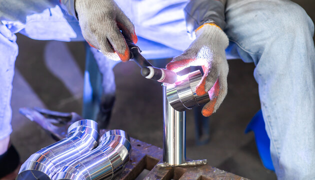 Selective focus to a worker performing precise TIG welding on stainless steel pipe parts in a factory workshop. Craftsmanship in metal fabrication and pipe welding process.
