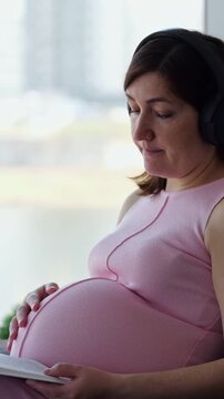 Serene Pregnant Lady Wears Headset Near Sunlit Window While Gently Caressing Bare Round Belly. Focused Expectant Parent Holds Document, Listening Carefully In Quiet Peaceful Indoor Environment.