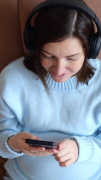 Expectant Mother Using Mobile Phone With Wireless Headphones Indoors, Passing Pregnancy Boredom In Cozy Home. Pregnant Woman Taps Smartphone Screen, Enjoying Digital Leisure While Resting.