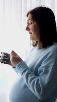 Pregnant Woman Swallowing Nutritional Supplement Pill Holds Textured Glass Of Water Near Window. Calm Expectant Mother Drinks Liquid Attentively, Maintaining Health Routine In Natural Daylight.