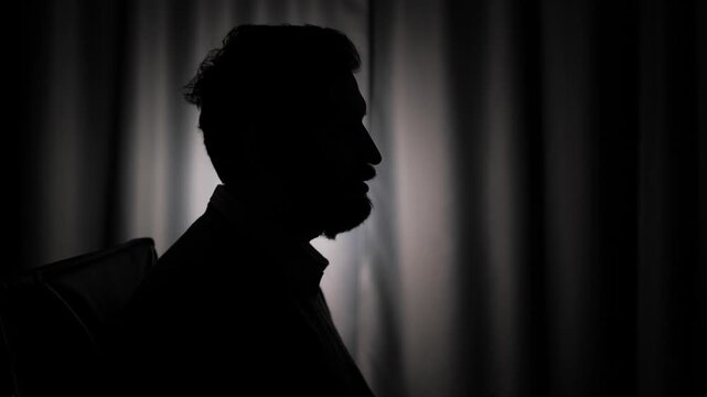 Hidden identity, unidentified person, silhouette, interview. Side view of dark male silhouette against light background. Man is speaking and looking at camera
