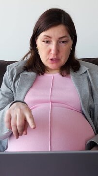 Pregnant Woman Participates In Virtual Meeting Sitting Relaxed On Sofa Indoors. Future Mother Gestures Gently While Discussing Maternity Advice With Focus During Calm Remote Communication.