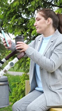 Young Mother Rests On Park Bench With Stroller While Checking Smartphone And Drinking Takeaway Coffee. Smiling Parent Enjoys Peaceful Outdoor Leisure During Pleasant Stroll With Baby Carriage.