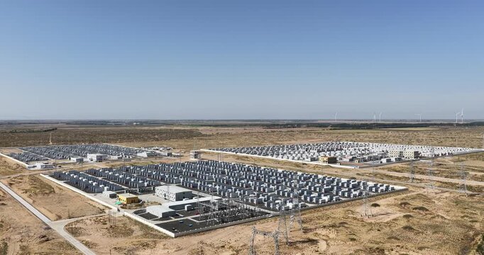Aerial drone footage orbiting an energy storage station in Inner Mongolia, rows of battery energy storage containers and a substation in sandy land under a clear blue sky, China.