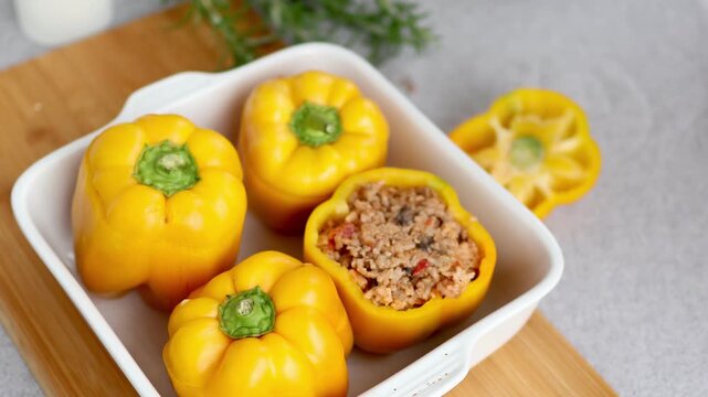 Woman sprinkles grated parmesan cheese over the filling of stuffed yellow bell peppers in a white baking dish before baking. Concept of traditional family recipes and homemade cooking.