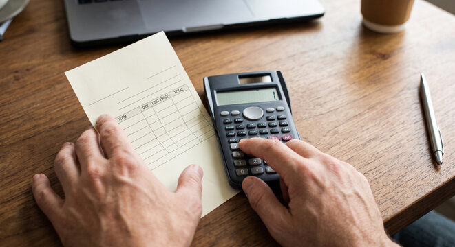 A person calculating expenses on a scientific calculator while holding an itemized receipt at a wooden desk with a laptop and pen