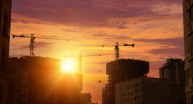 Construction site with tower cranes and modern high-rise building silhouettes against bright orange sunset sky in urban area dusk.