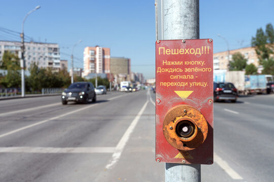 Close-up view of pedestrian push button on pole with Russian instruction sign along busy city street with traffic in background.