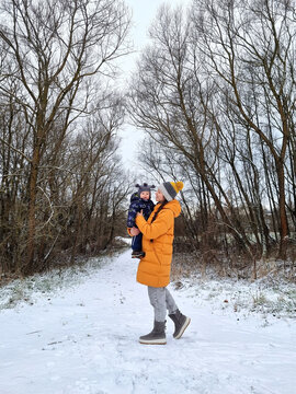 Vertical full length shot of young mother in bright yellow down jacket holding baby toddler standing on snowy path in winter forest park with bare trees under overcast sky daylight lifestyle