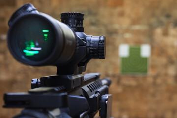 Close up of optical rifle scope with elevation turrets mounted on firearm pointing towards blurred paper target inside indoor shooting range © Dmitri