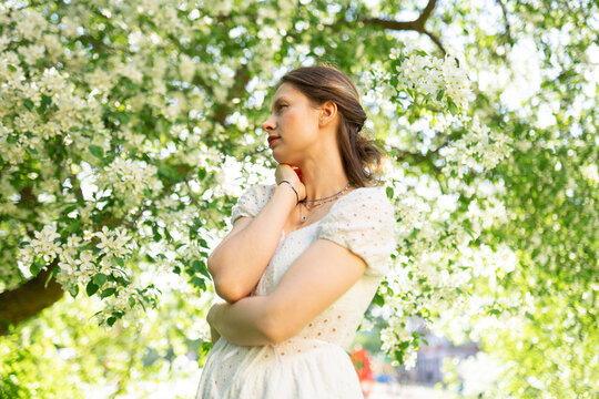 A woman in a white dress poses by a blooming apple tree