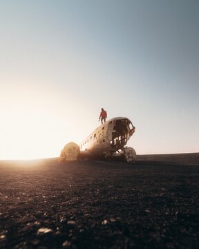 View of a traveler in a red jacket on the Solheimasandur plane wreck against black volcanic sand during golden hour in Skeidflotur, Myrdalshreppur, Iceland.