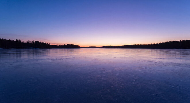 Peaceful winter lake with snowy shore reflecting soft twilight colors at sunset under clear evening sky