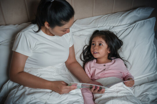 happy mother read story in the book for preschool child girl in bed at night