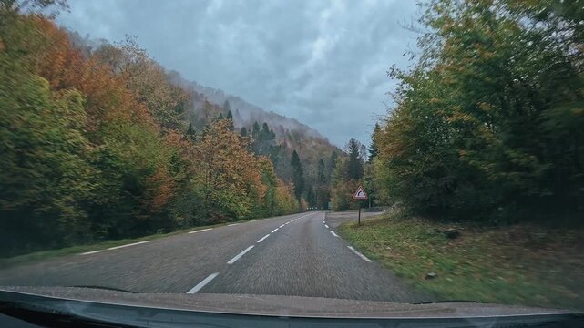 Route de montagne sinueuse traversant une for&ecirc;t aux couleurs automnales, envelopp&eacute;e de brume sous un ciel couvert. Une atmosph&egrave;re calme et myst&eacute;rieuse au c&oelig;ur d&rsquo;un paysage naturel de montagne.