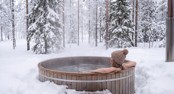 A woman relaxing in an outdoor wooden hot tub bath surrounded by winter snow and forest trees