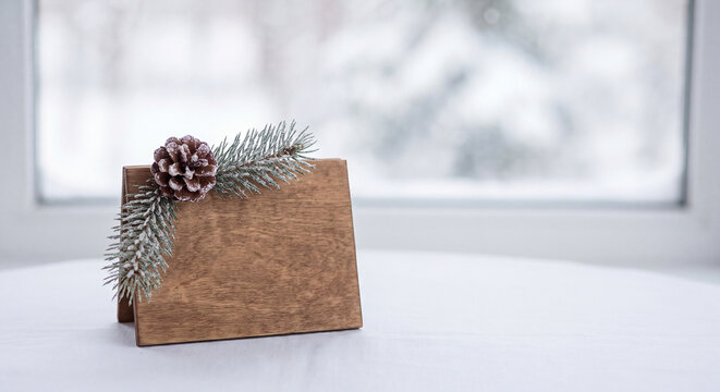 A rustic wooden place card holder decorated with a pine cone rests on a bright windowsill