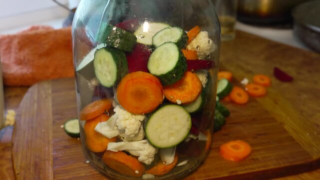 Organic produce preservation with Cucumis sativus in home kitchen. Static close-up shot showing hand adding sliced vegetables into clear glass storage jar.