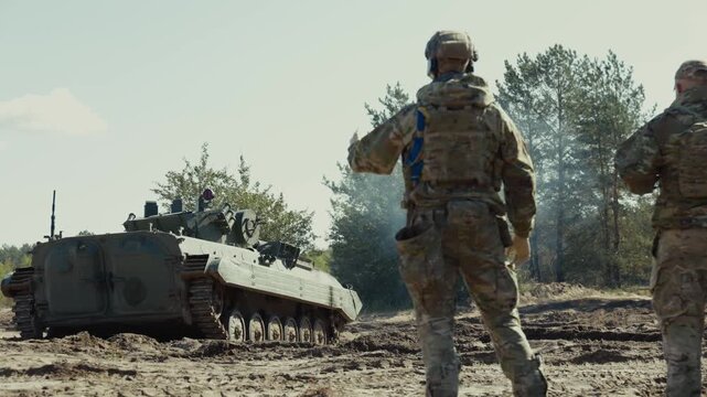 A soldier raises his hand to signal direction to an approaching armored tracked vehicle on a sandy training field.