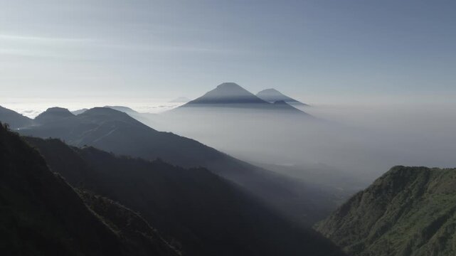Hiker Standing on Mountain Ridge Overlooking Volcanic Peaks Emerging from Morning Mist at Mount Bismo, Central Java, Indonesia.