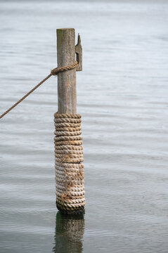 Holzpfahl mit aufgewickeltem Schiffstau steht im ruhigen Wasser eines Hafens bei weichem, bedecktem Tageslicht in maritimer, stilvoller Atmosph&auml;re.