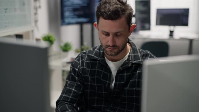 Caucasian male programmer using a mobile phone while sitting at a computer workstation in a modern office, communication and software development workplace concept.