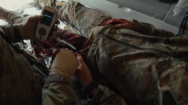 A gloved medic attaches a pulse oximeter clip to monitor oxygen levels on a wounded soldier lying on a stretcher.