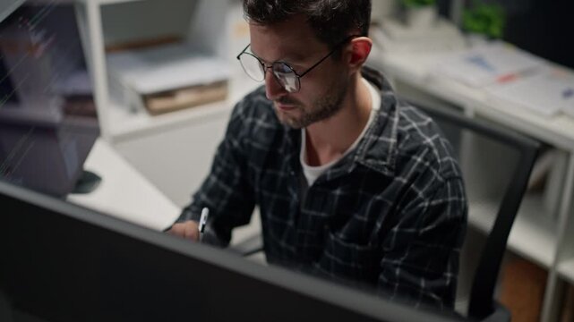 Caucasian male programmer wearing glasses and using a stylus with tablet while working at a computer workstation in a modern office, software development and technology workplace concept.