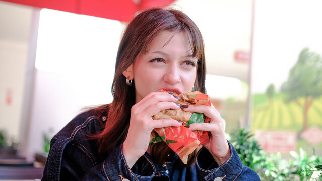 Young woman taking a big bite of a burger with lettuce and tomato at an outdoor cafe, holding it with both hands, long brown hair, dark denim jacket, red umbrella above, bright day, fast food