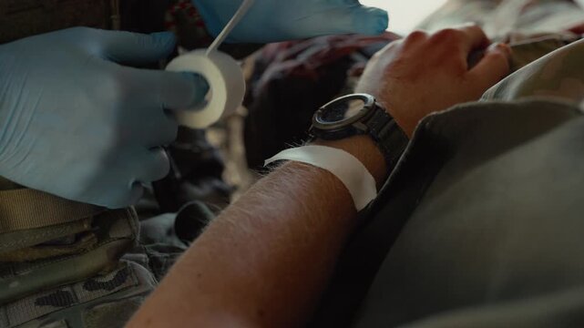Gloved military medics apply white tape to a patient's wrist inside a vehicle, preparing the arm for further treatment, trauma shears nearby.