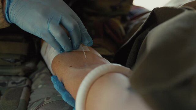 A gloved medic positions an IV needle above a patient's tourniqueted arm, preparing for insertion at a tactical medical point.