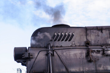 Dark front section of a black steam locomotive releasing a plume of grey smoke into a pale sky, industrial railway background © syntheticmessiah