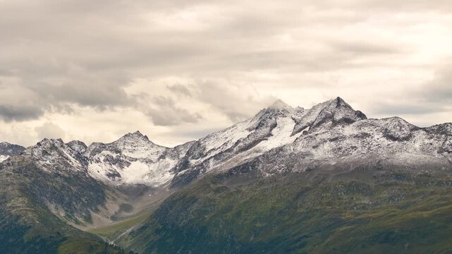 Slow aerial pan across jagged snow dusted peaks and a rugged scree valley near Gletsch, Switzerland, beneath an overcast sky with warm gray clouds.