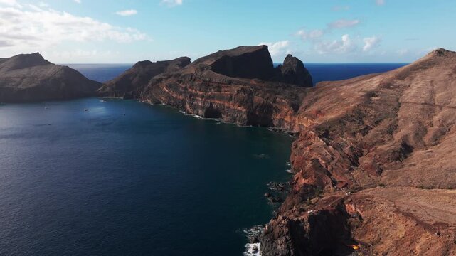 Aerial glide beside orange and dark layered cliffs at Ponta de Sao Lourenco, Madeira, overlooking sailboats in a blue bay, breaking waves and a path across the dry headland.