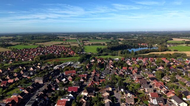 Small German town with dense red-tiled rooftops spreads beside calm reflective lake and green farmland, wooded hills and open fields stretching under wispy autumn sky. Aerial drone lateral wide shot