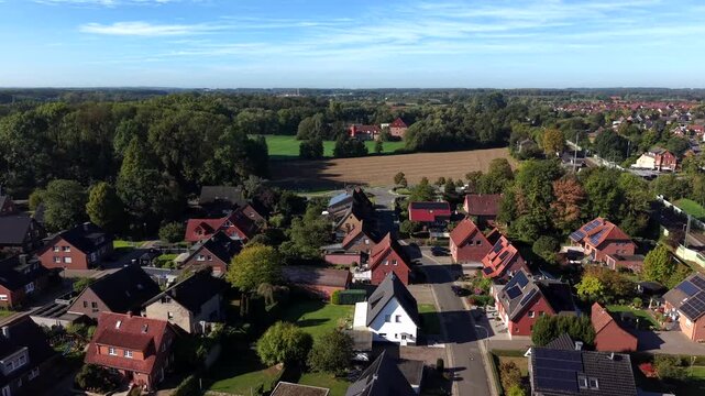 Red-roofed single-family houses with solar panels line quiet German suburban street bordering golden farmland and dense trees, passing train visible in distant background. Aerial drone flyover shot