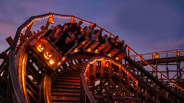 Motion blur riders in pumpkin-decorated coaster car climbing wooden track lit with string lights