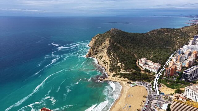 Rocky forested headland meets turquoise Mediterranean Sea with breaking waves, a sandy beach and high-rise hotel buildings along the coast of Benidorm, Costa Blanca, Spain
