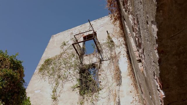 Weathered hacienda wall featuring a rusted metal balcony and climbing vines across cracked plaster. Blue sky highlights the deteriorating colonial architecture and aged exterior in Mexico.