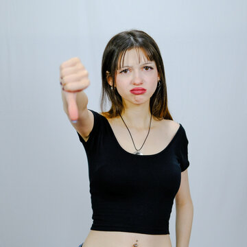 Young woman showing thumbs down gesture with disapproval in studio on light gray background, dislike, failure, rejection, negative feedback, bad review, disagreement concept, portrait, copy space