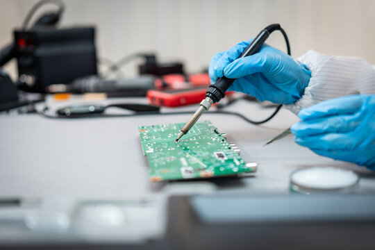 Close-up of a technician wearing blue gloves soldering a green circuit board at an electronics repair workbench.