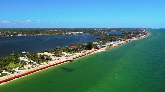 An aerial view follows a narrow residential shoreline, with a coastal road and seawall between green open water and a darker bay.