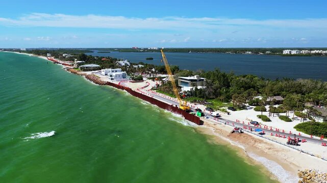 A yellow crane stands beside a long steel sheet-pile wall along a sandy shoreline, with traffic barriers, houses and a broad waterway behind the road.