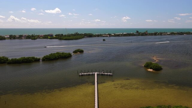 A T-shaped dock extends into shallow lagoon water as small boats leave wakes beyond mangrove islets, with the ocean visible past the distant shore.