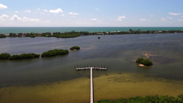A narrow pier ends in a T-shaped platform in a shallow lagoon, with mangrove islets and a distant shoreline lined with buildings.