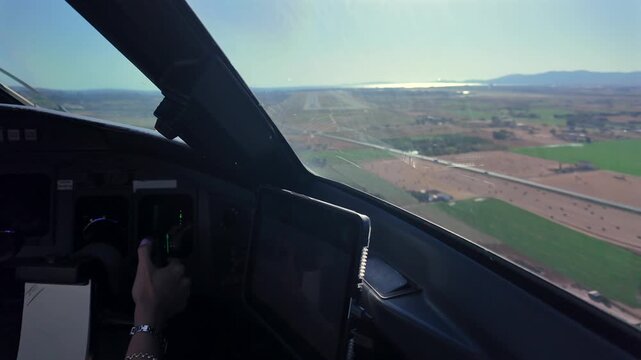 Hand of a young female pilot with pink painted nails on the flight controls of a modern jet cockpit in a bumpy approach to coastal Palma de Mallorca airport runway with a blinding sun ahead.