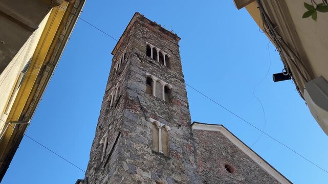 Low-angle view of the Romanesque stone bell tower and facade of the Pieve di Sant'Andrea in Sarzana, Liguria, under a clear blue sky.