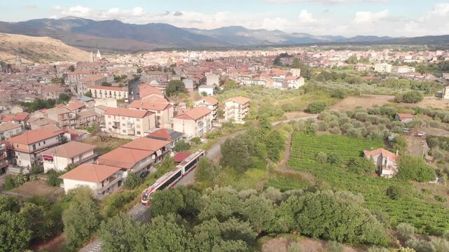 Aerial drone shot following the Ferrovia Circumetnea train as it enters the town of Randazzo in Sicily, with houses and the surrounding volcanic landscape visible.