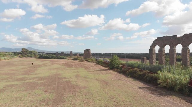 Aerial drone tracking shot flying parallel to the railway as a train passes through the Parco degli Acquedotti in Rome, with ancient aqueducts visible nearby.