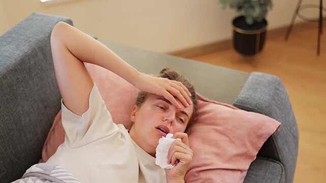 A woman lies on a couch with a tissue in her hand. She has a hand on her forehead and appears to be unwell. Soft light comes through the window